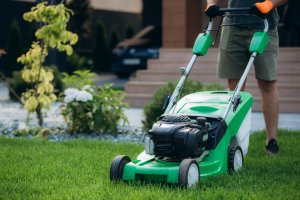 A young man is mowing a lawn with a lawn mower in his beautiful green floral summer garden.